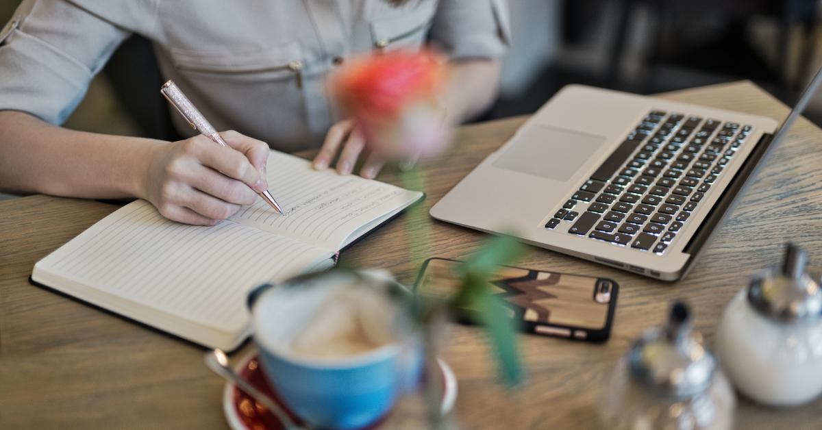 man writing in notebook next to laptop and cup of coffee
